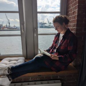 Frau auf Fensterbank sitzend mit Buch und im Hintergrund der Hafen in Hamburg