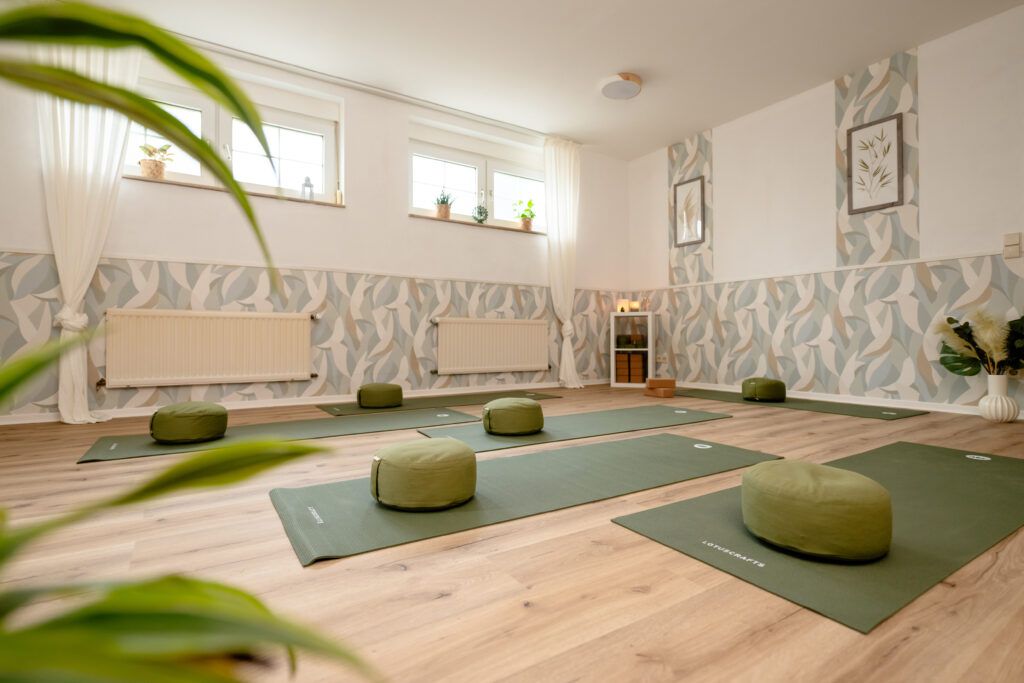 Yoga-Raum mit Matten auf dem Boden und Meditationskissen darauf. Im Hintergrund brennen Kerzen und es sind noch Blöcke und Yoga-Gurte zu sehen.