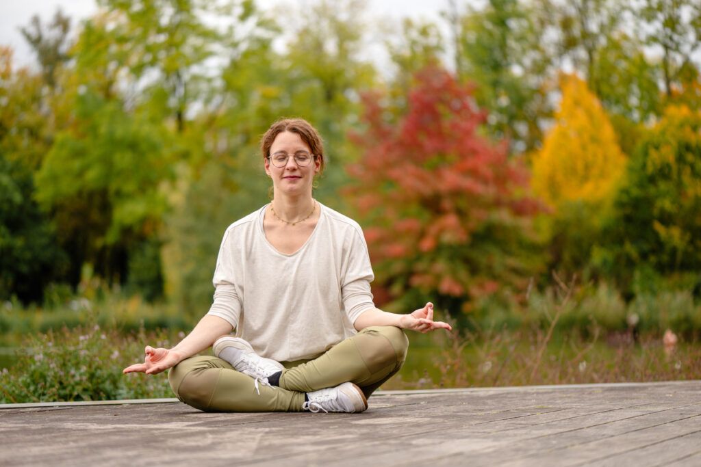 Frau sitzt im halben Lotussitz auf einer Terrasse. Hinter sich ein See. Sie hat die Hände in Meditationshaltung und die Augen geschlossen. 
